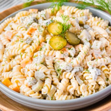 square image of dill pickle pasta salad in a serving bowl with pickles and fresh dill on top
