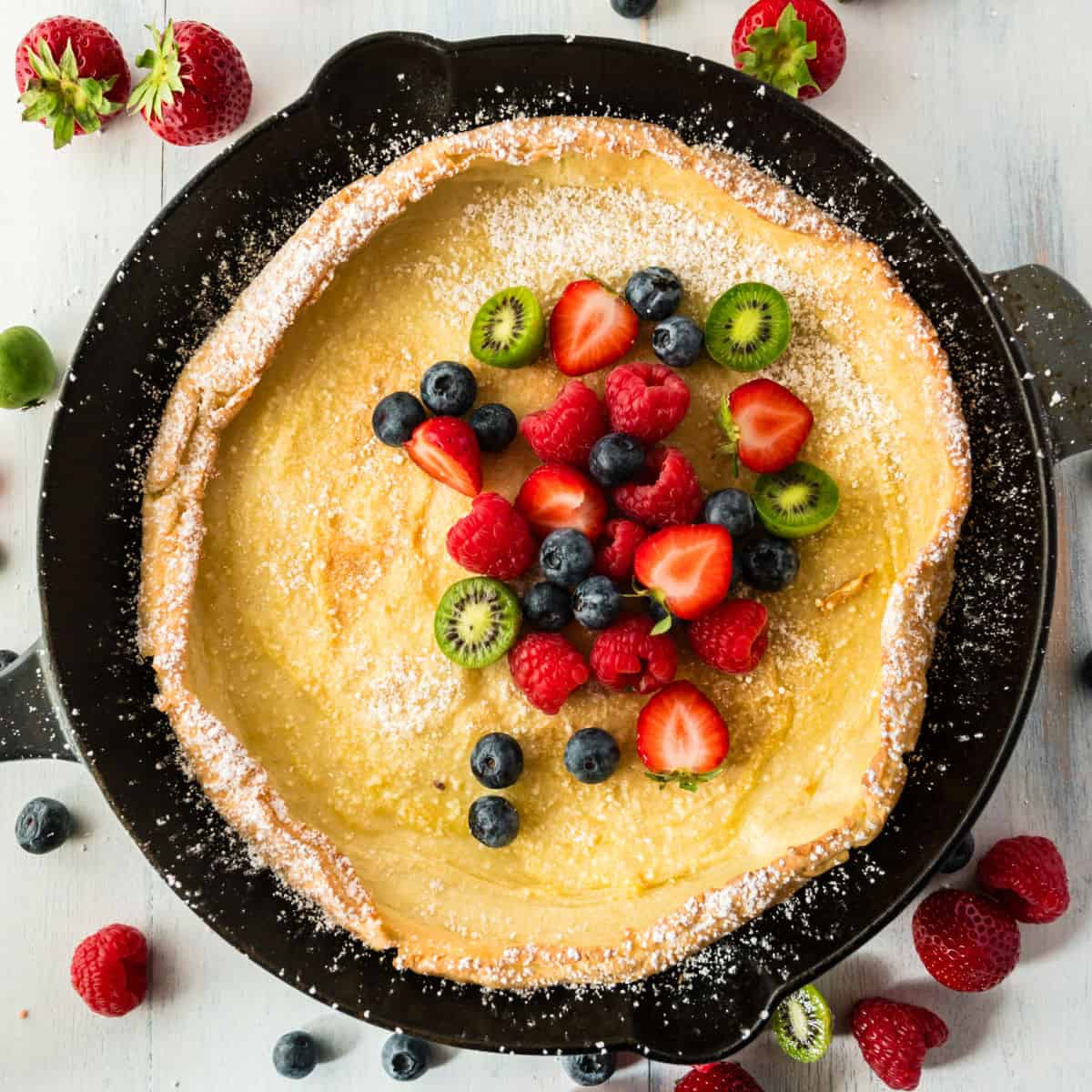 square image of a dutch baby topped with powdered sugar and fresh fruit in a cast iron skillet