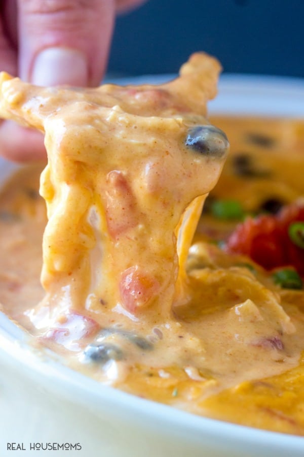 Tortilla chip lifting a bite of easy chicken queso dip from the bowl with gooey cheese dripping down