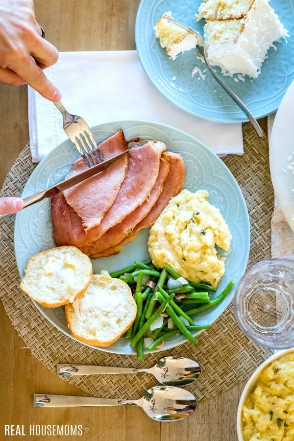 persona cutting into ham slices on a dinner plate with other side dishes