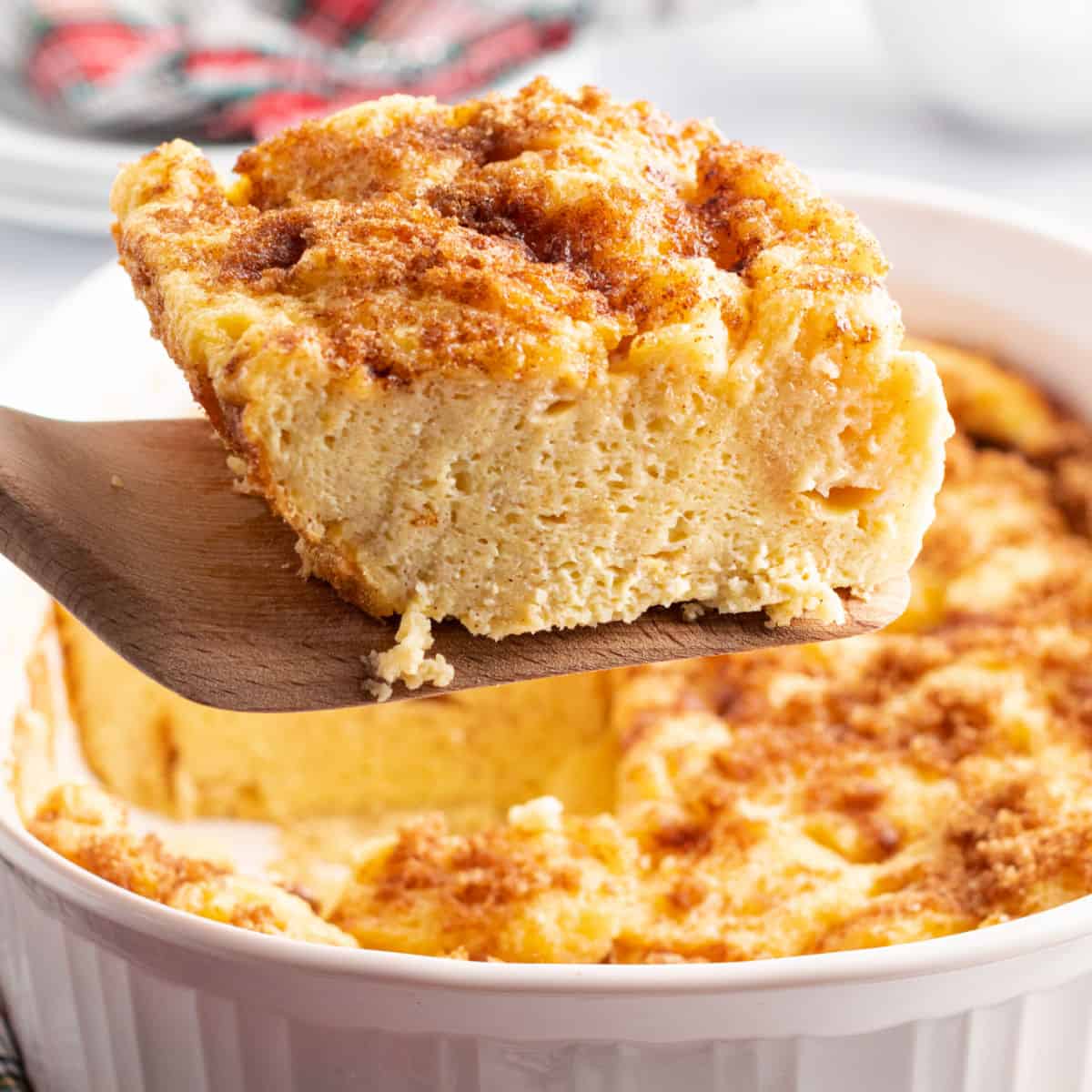 square image of a slice of eggnog french toast bake on a wooden spatula over the baking dish