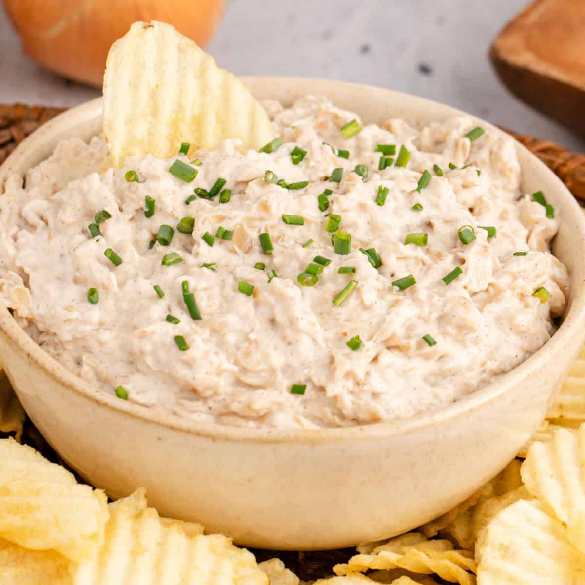 square image of french onion dip in a serving bowl with a chip stuck into the dip