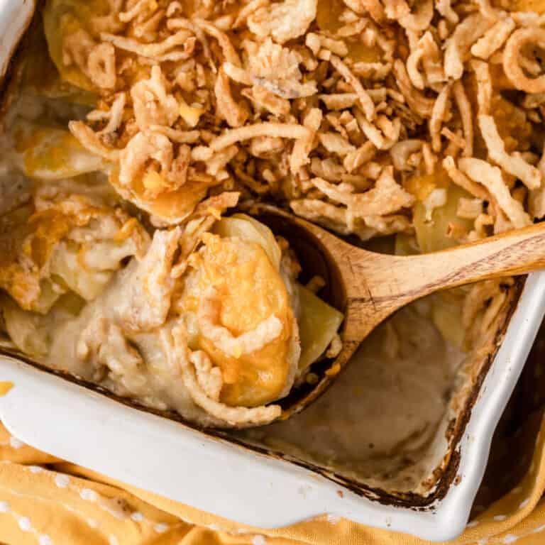 close up of French Onion Scalloped Potatoes in a white pan with a wooden spoon full of a scoop of potatoes