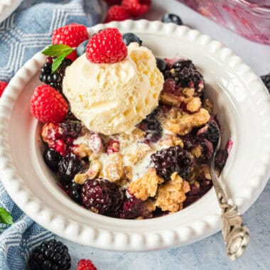 square image of fresh berry crisp in a bowl with ice cream and berries