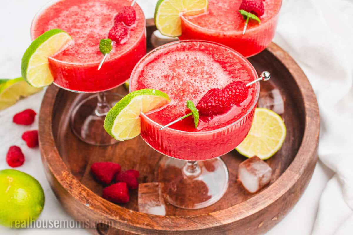 three frozen raspberry margaritas on a wooden serving tray with fresh fruit