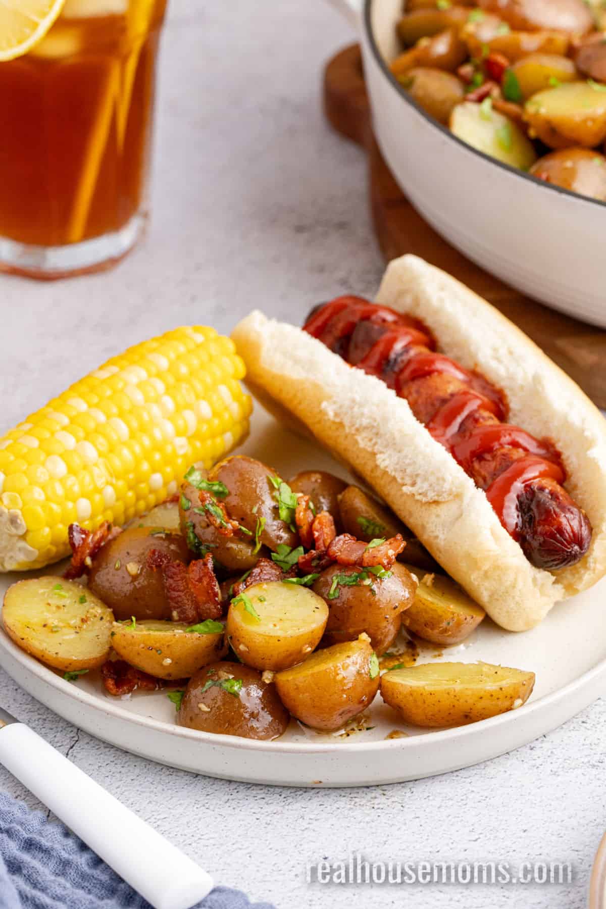 serving of german potato salad with an ear of corn and a hot dog with ketchup on a white plate