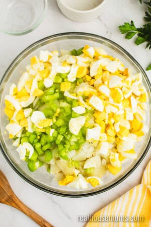 chopped eggs, cooked potatoes, chopped celery, and onion added to a large glass bowl with potato salad dressing