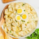 square image of grandma's best potato salad with hard boiled egg slices on top in a serving bowl with a wooden spoon