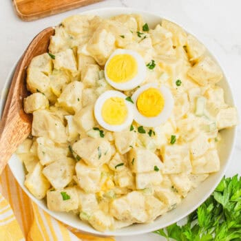 square image of grandma's best potato salad with hard boiled egg slices on top in a serving bowl with a wooden spoon