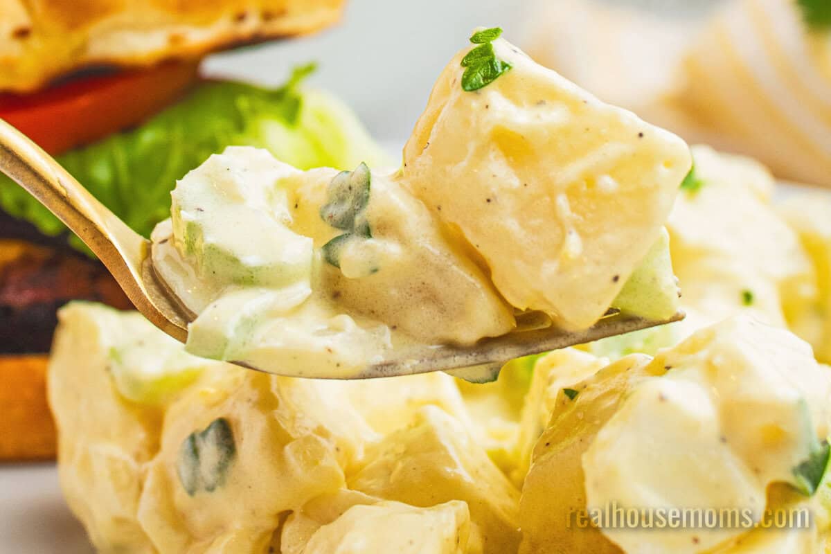bite of grandma's best potato salad on a fork over a plate with a burger and potato salad