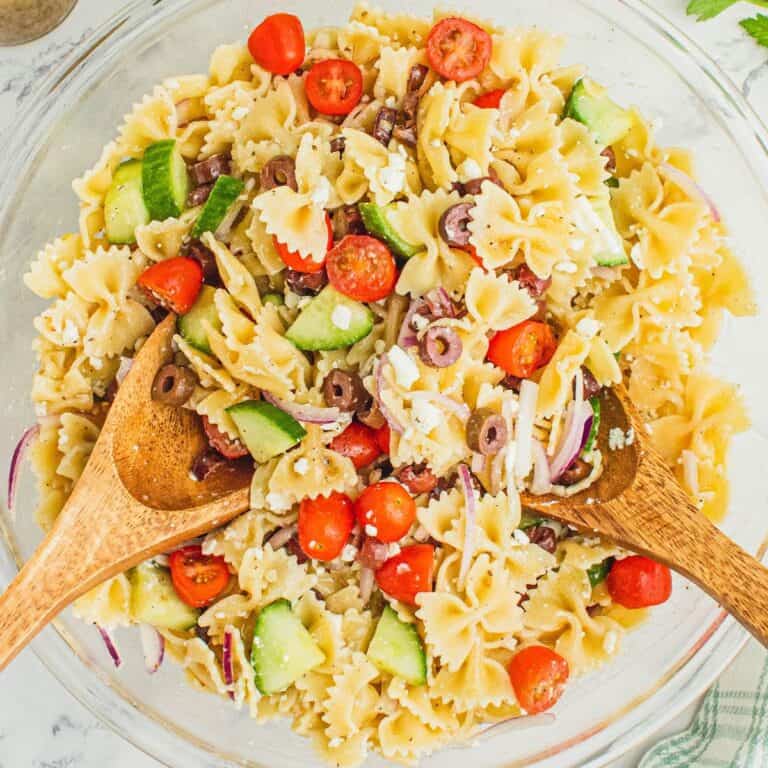 square image of greek pasta salad in a serving bowl with wooden spoons