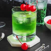 square image of a green goblin cocktail in a rocks glass garnished with maraschino cherries on a cocktail pick