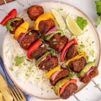 square image of grilled chicken fajita skewers with chopped cilantro and lime wedges served over rice on a plate