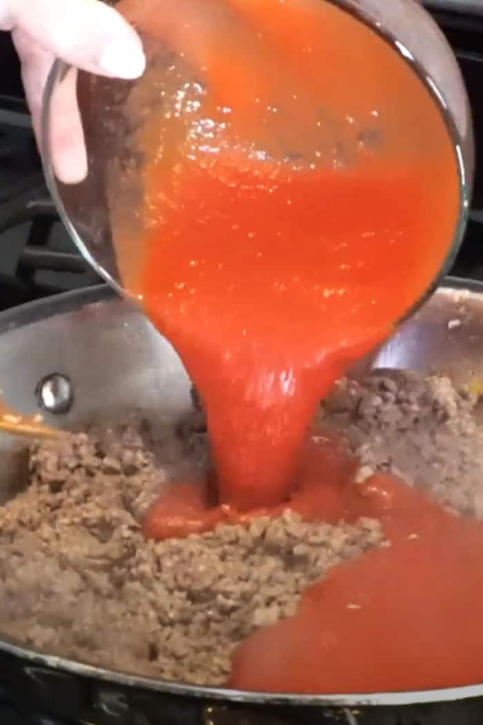tomato sauce being poured into cooked ground beef in a skillet