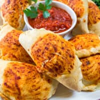 square image of ham & cheese mini calzones piled on a plate with a bowl of marinara for dipping