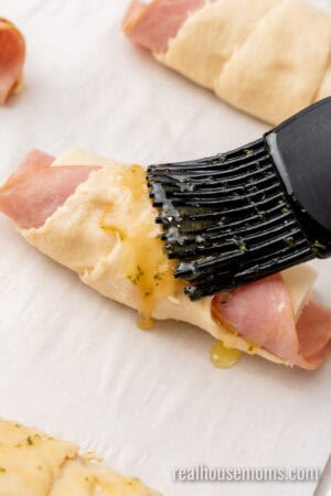 butter and brown sugar glaze being brushed onto uncooked ham and cheese crescent rolls