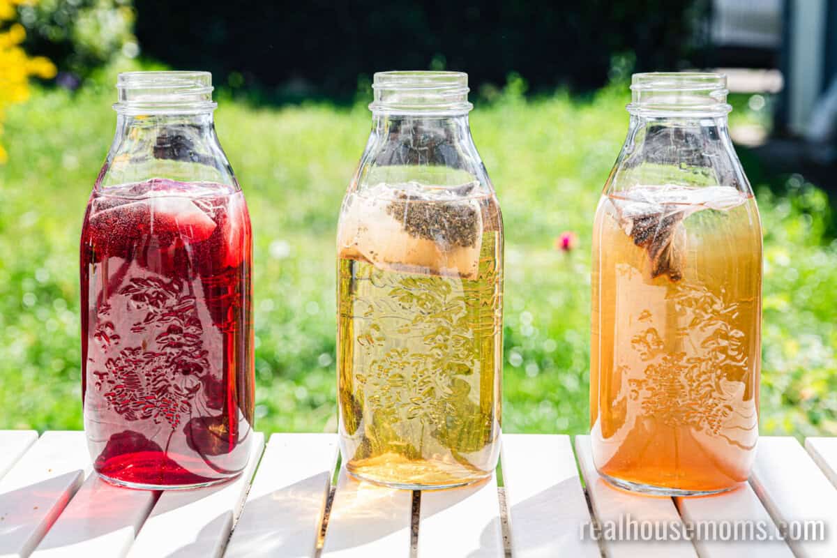 three glass jars on a porch railing, each with a different type of tea bag steeping inside