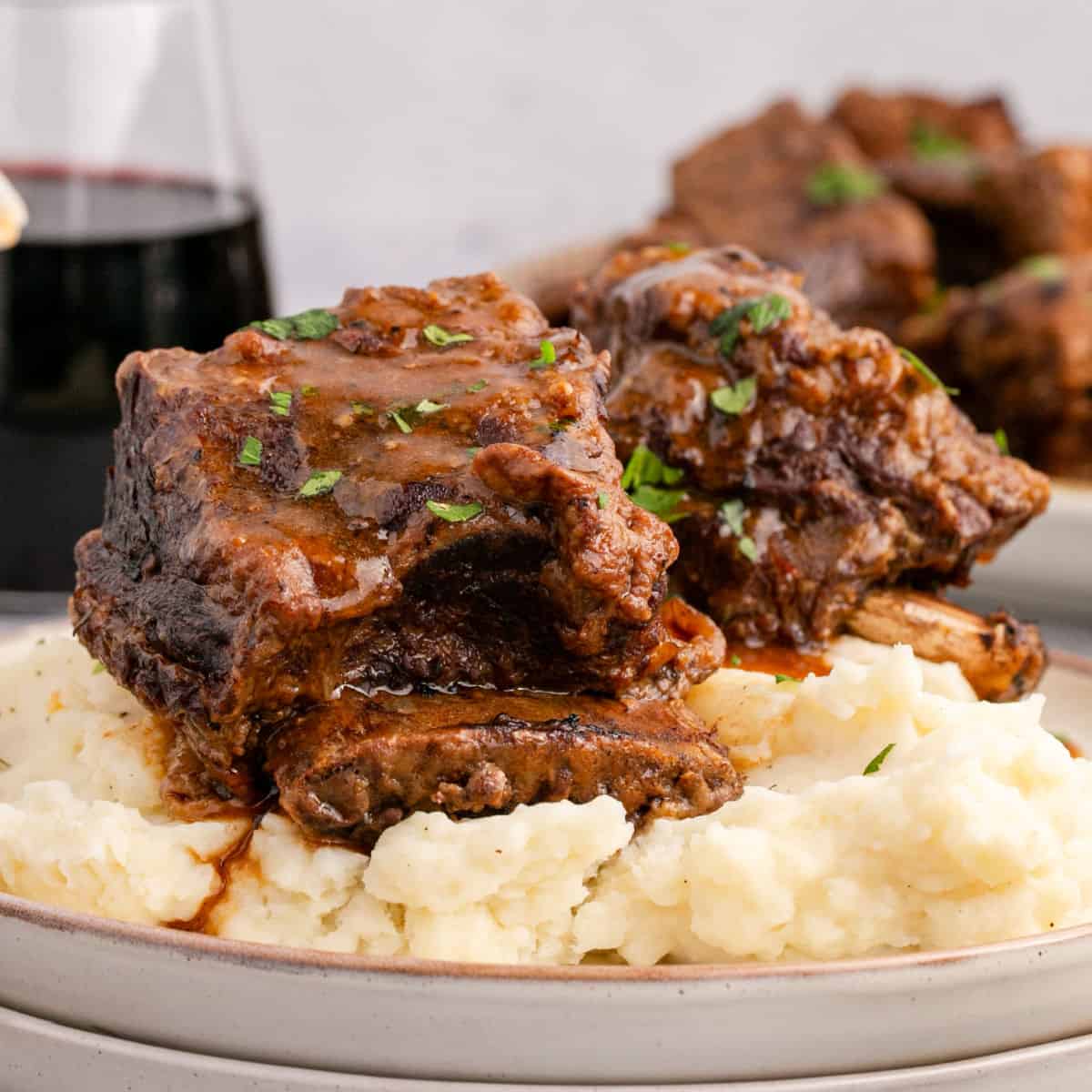 square image of instant pot short ribs with gravy plated over mashed potatoes next to a glass of red wine