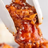 close up of a korean fried chicken piece with sesame seeds being picked up by tongs with the tite of the post on top of the image in bluena and black lettering