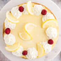 sqiare image of lemon cheesecake topped with whipped cream dollops, lemon slices, and fresh raspberries