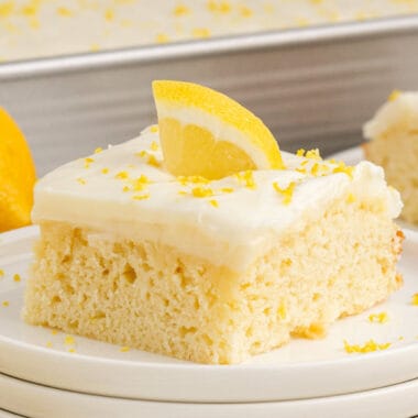 square image of a slice of lemon poke cake topped with lemon zest and a lemon slice on a plate
