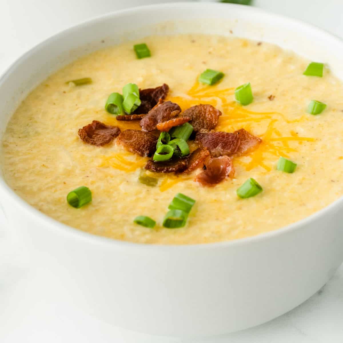 square image of cauliflower chowder in a white bowl topped with cheese, bacon, and green onion
