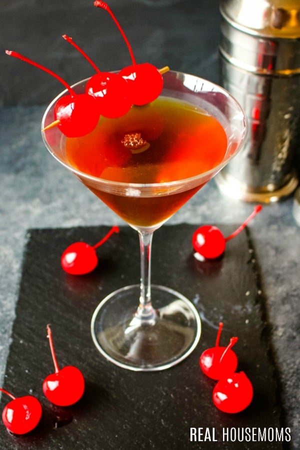 Close up of manhattan cockail in a martini glass garnished with cherries alongside a cocktail shaker
