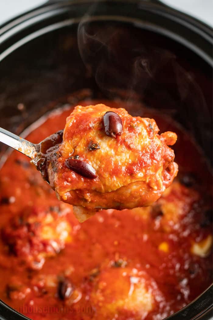 serving spoon lifting a chicken thigh covered in tomato sauce and olives out of a crock pot