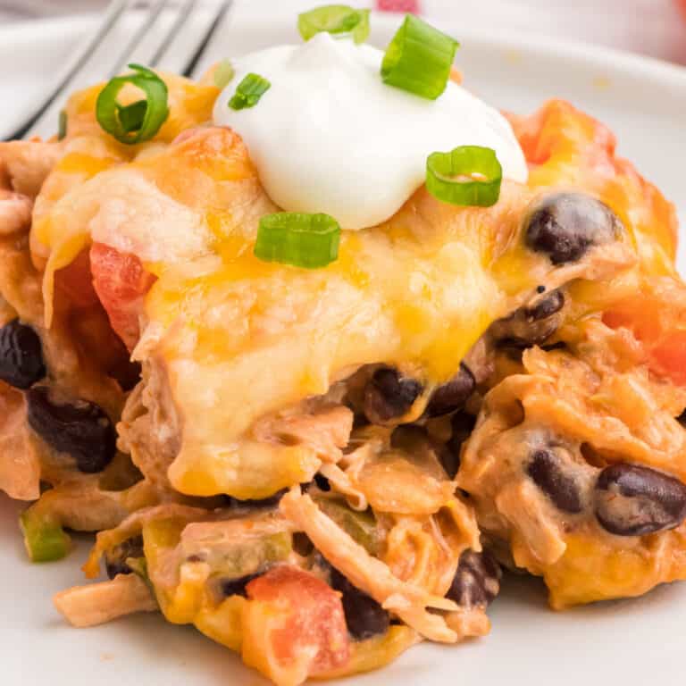 square image of a slice of mexican chicken casserole on a plate with sour cream and green onions on top
