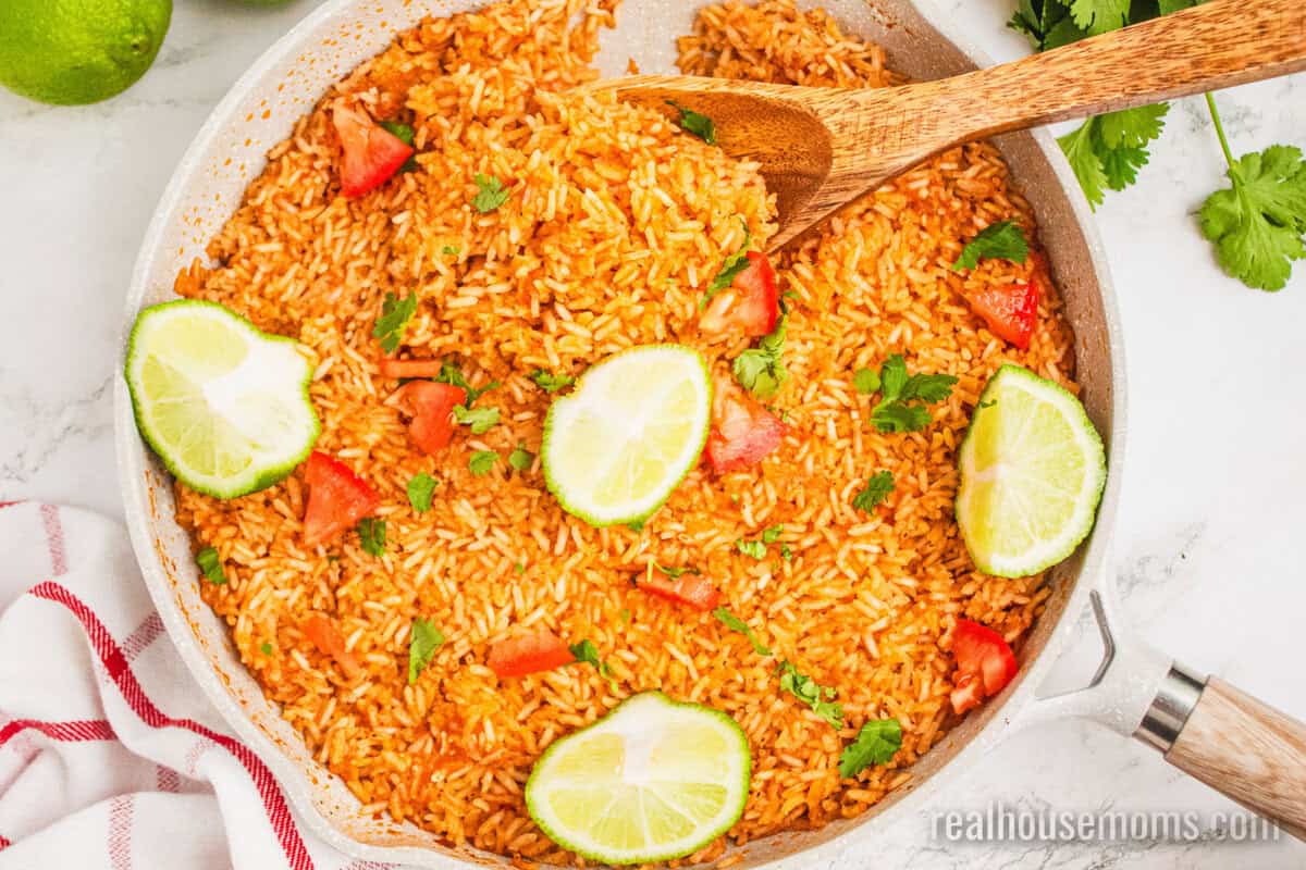 wooden spoon resting in a skillet of mexican rice topped with diced tomato, cilantro, and lime wedges