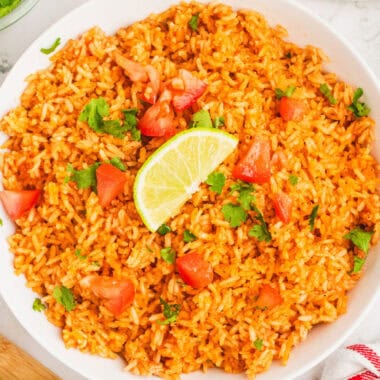 square image of mexican rice topped with cilantro, tomatoes, and a lime wedge in a white serving bowl