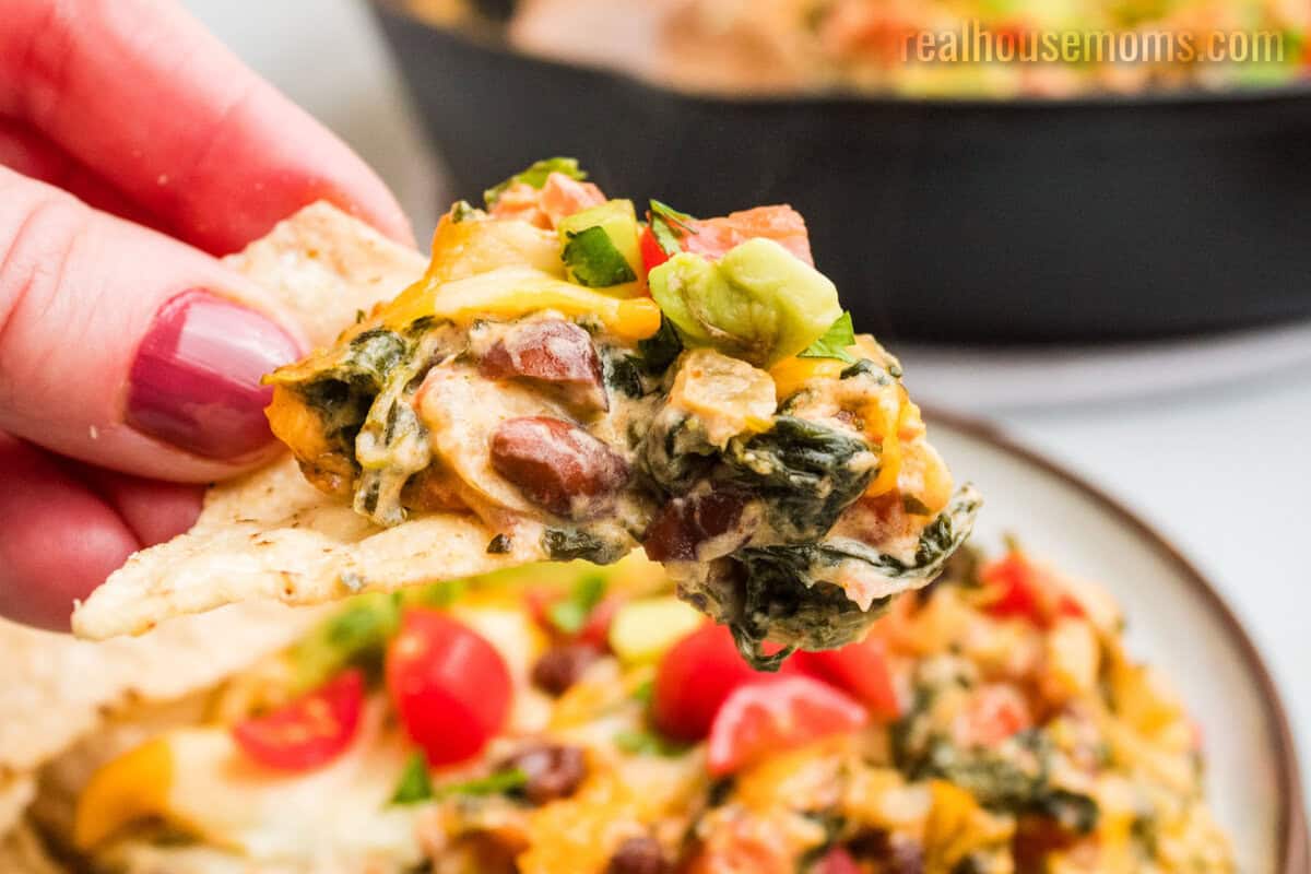 hand holding a tortilla chips loaded with mexican spinach dip over a plate