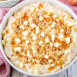 square image of apple cheesecake dip in a shallow bowl garnished with caramel, nuts, and apples