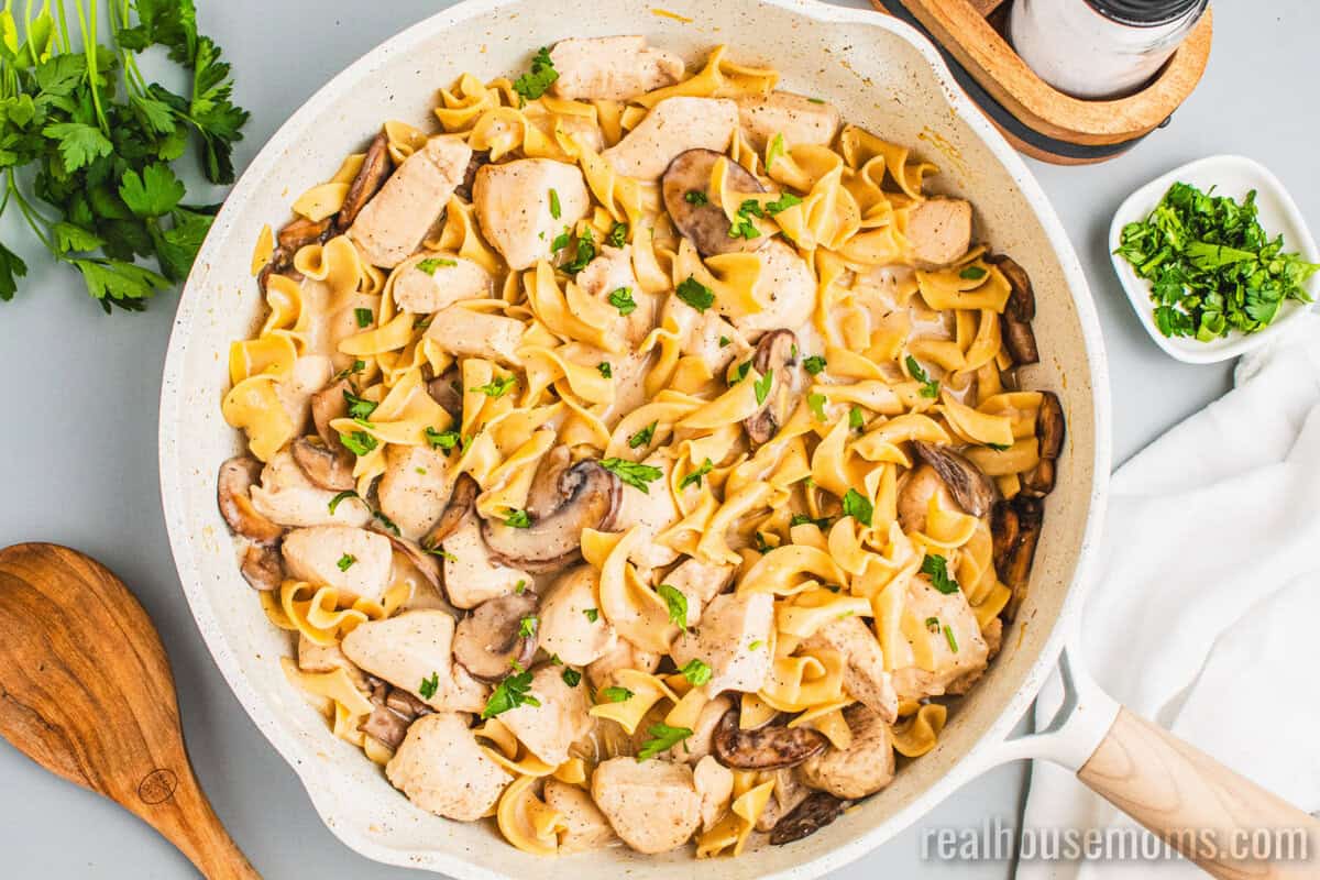 skillet of chicken stroganoff next to a wooden serving spoon and a bowl of chopped parsley