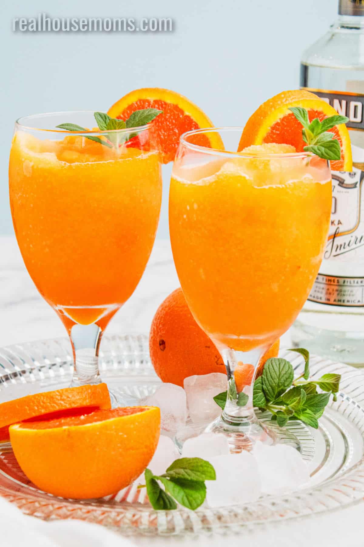 two orange vodka slushes on a tray next to a bottle of vodka