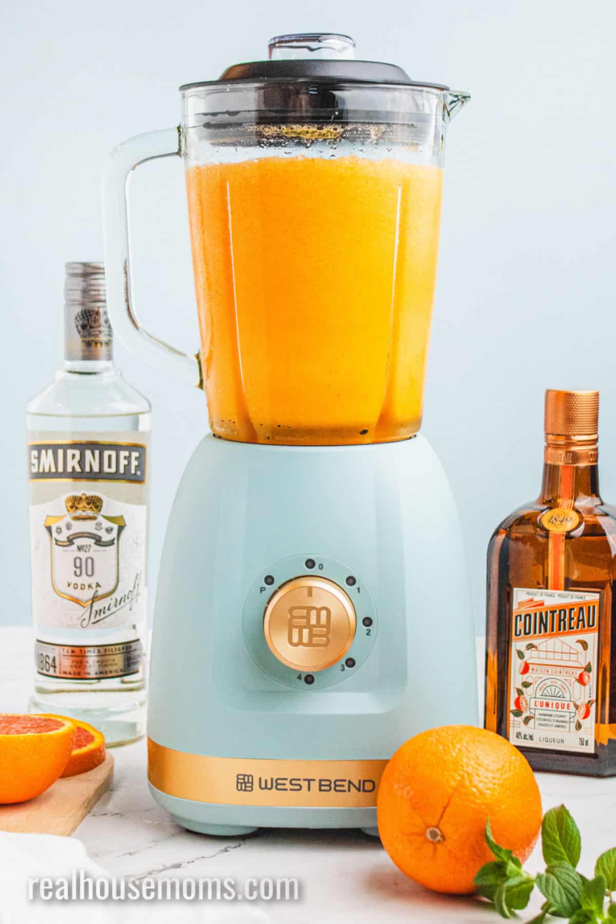 blender of orange vodka slush next to a bottle of vodka, Cointreau, and oranges