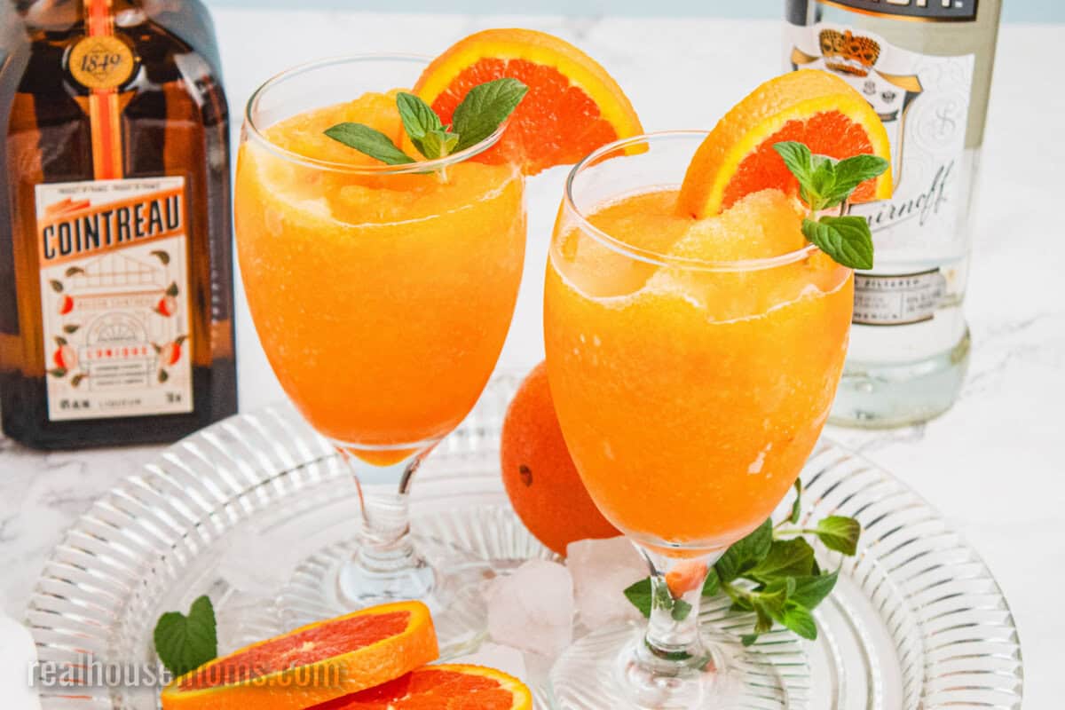 two orange vodka slushes garnish with a half orange slice and a mint spring on a tray next to bottles of vodka and cointreau