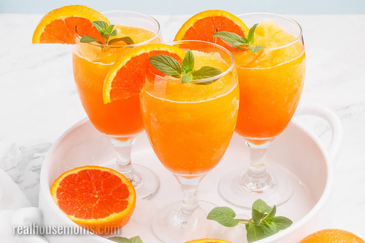 three glasses or orange vodka slush on a white tray with an orange half and mint