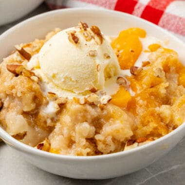 square image of a bowl of peach dump cake topped with ice cream and chopped nuts