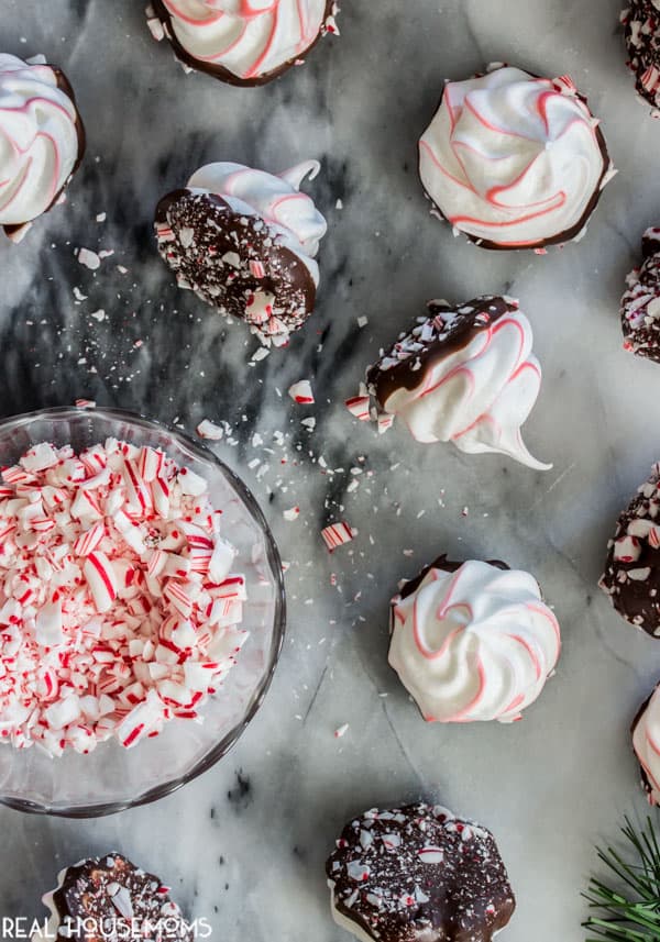 Peppermint Bark Meringues on a counter top, dipped in chocolate ready to be dipped into a bowl of crushed candy canes