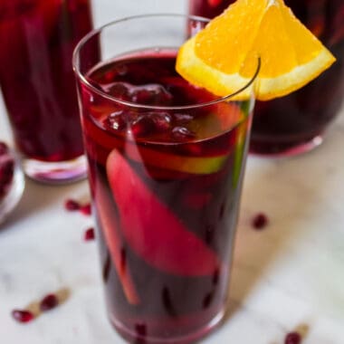 square image of pomegranate sangria in a tall glass with orange wedge and pomegranate arils