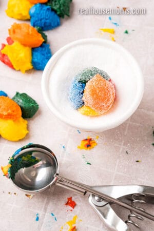 ball of rainbow cake mix cookie dough rolled in a bowl of sugar next to a cookie scoop