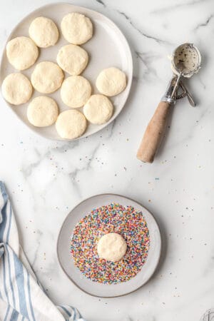 flatten cookie dough balls being dipped into sprinkles on a plate next to the cookie scoop