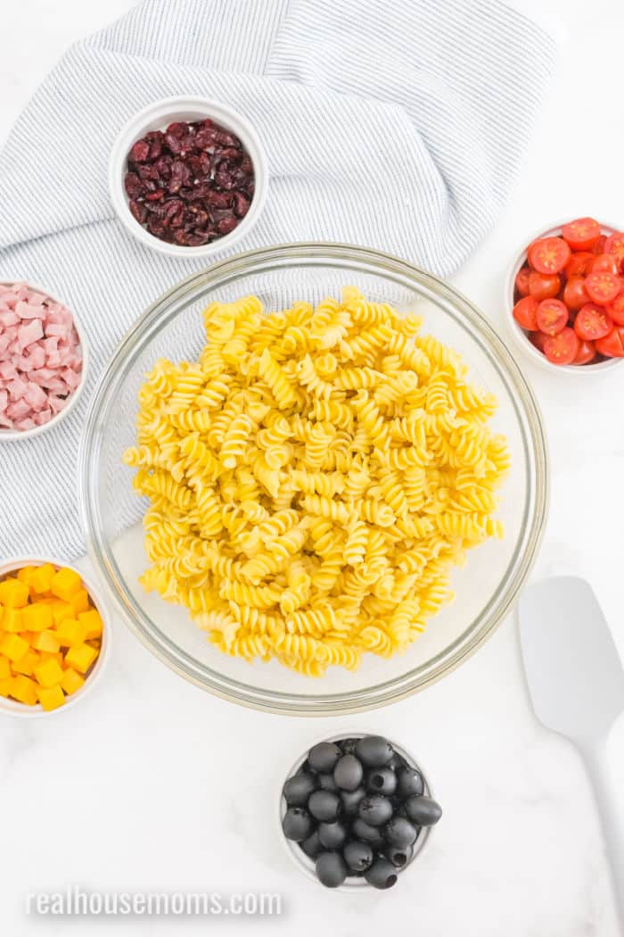 cooked rotini pasta in a bowl, next to bowls of tomatoes, raisins, cheese, ham, and olives