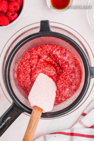 cooked raspberries being pressing through a mesh sieve with a rubber spatula into a glass bowl