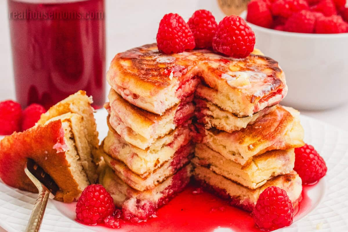 Pancakes topped with raspberry syrup and fresh raspberries next to a bite of pancakes on a fork