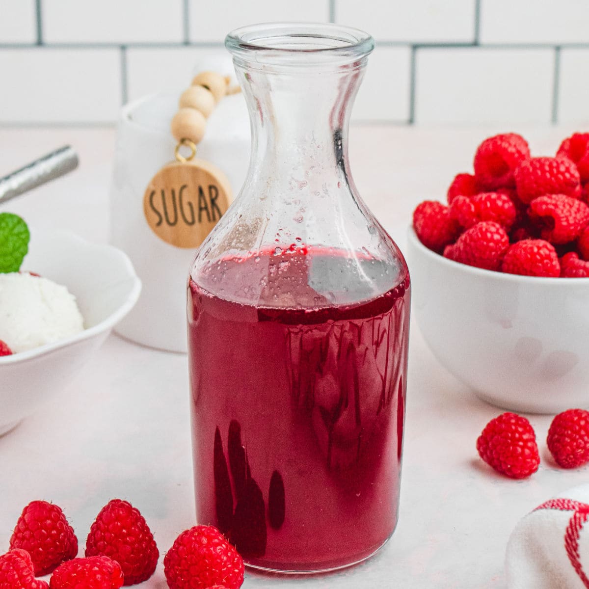 square image of a glass bottle of raspberry syrup next to a bowl of raspberries and a bowl of vanilla ice cream