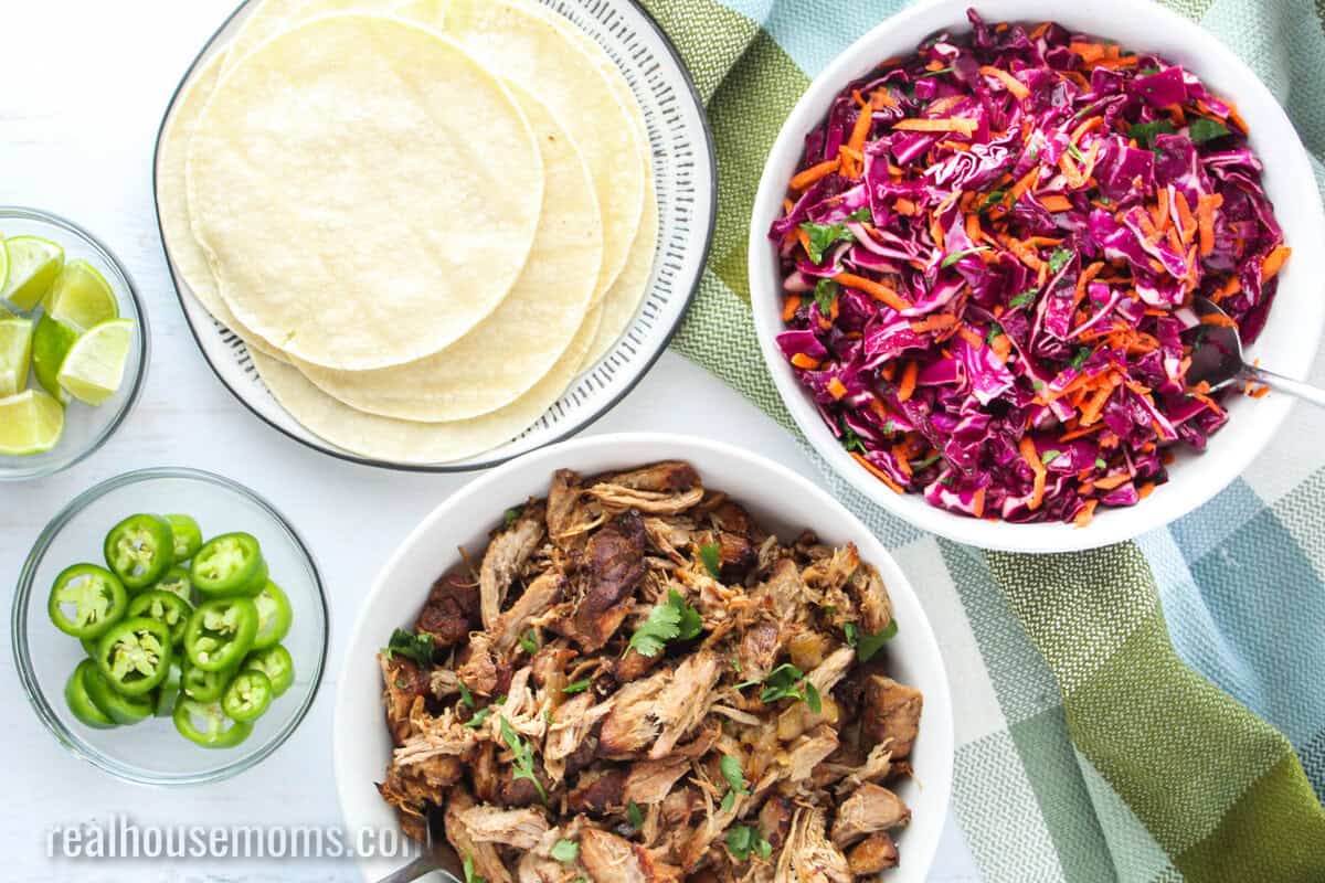 bowl f red cabbage slaw next to tortillas, carnitas, sliced jalapeno, and limes for tacos
