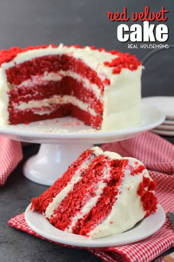 A slice of 3-layer red velvet cake on a dessert plate with the rest of the cake on a cake stand behind it