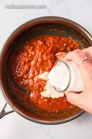 coconut milk being poured into a skillet with curry pasted, diced tomatoes, garlic, and ginger